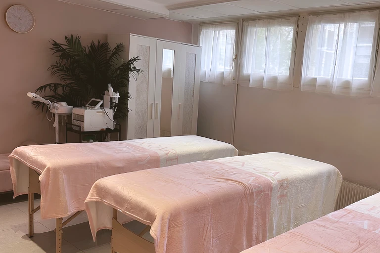 Salle de soins esthétiques avec plusieurs tables de massage recouvertes de draps roses et blancs, une armoire blanche et du matériel de soin.