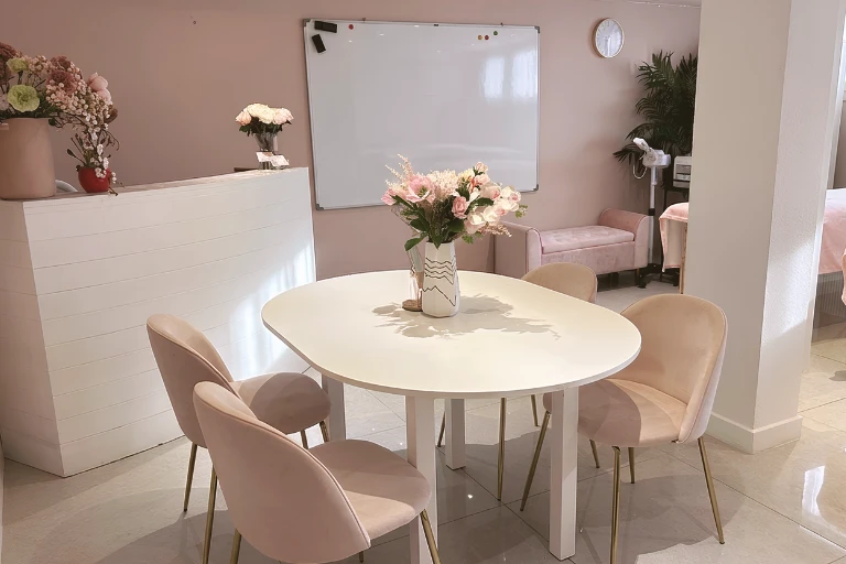 Une salle d'attente rose avec une table blanche ovale entourée de quatre fauteuils en velours rose, une réception et un tableau blanc au mur.